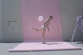 Man performing modern dance in a bathing suit against a pink background outside with a pink balloon tied to his foot.